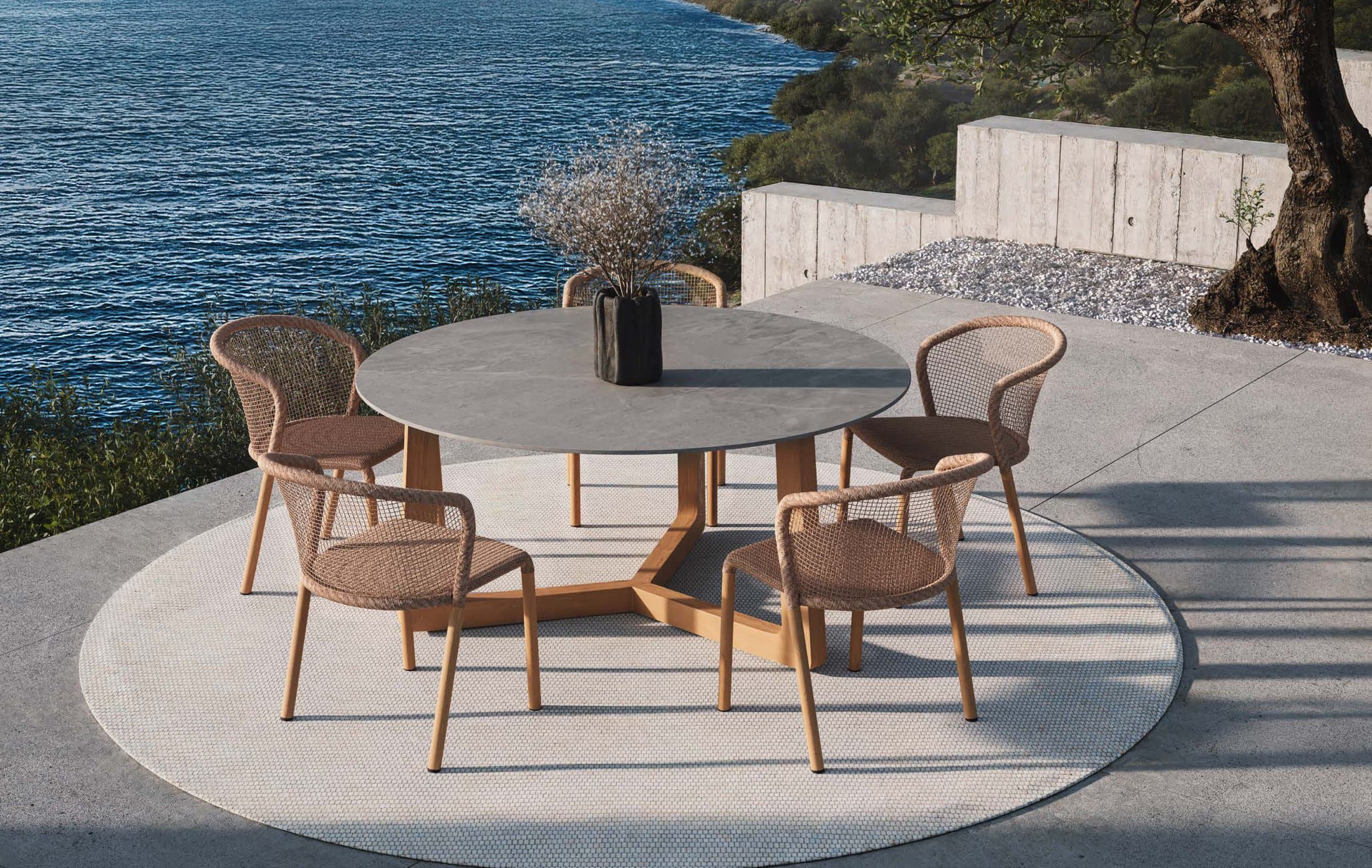 Drew round dining table with woven arm chairs on a terrace above the sea.