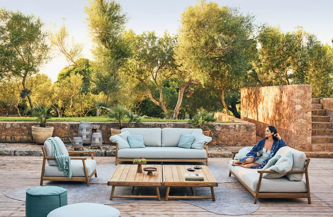 Manutti Muyu teak outdoor sofa, lounge chairs and low tables on a timber deck among olive trees, beside a stone wall.
