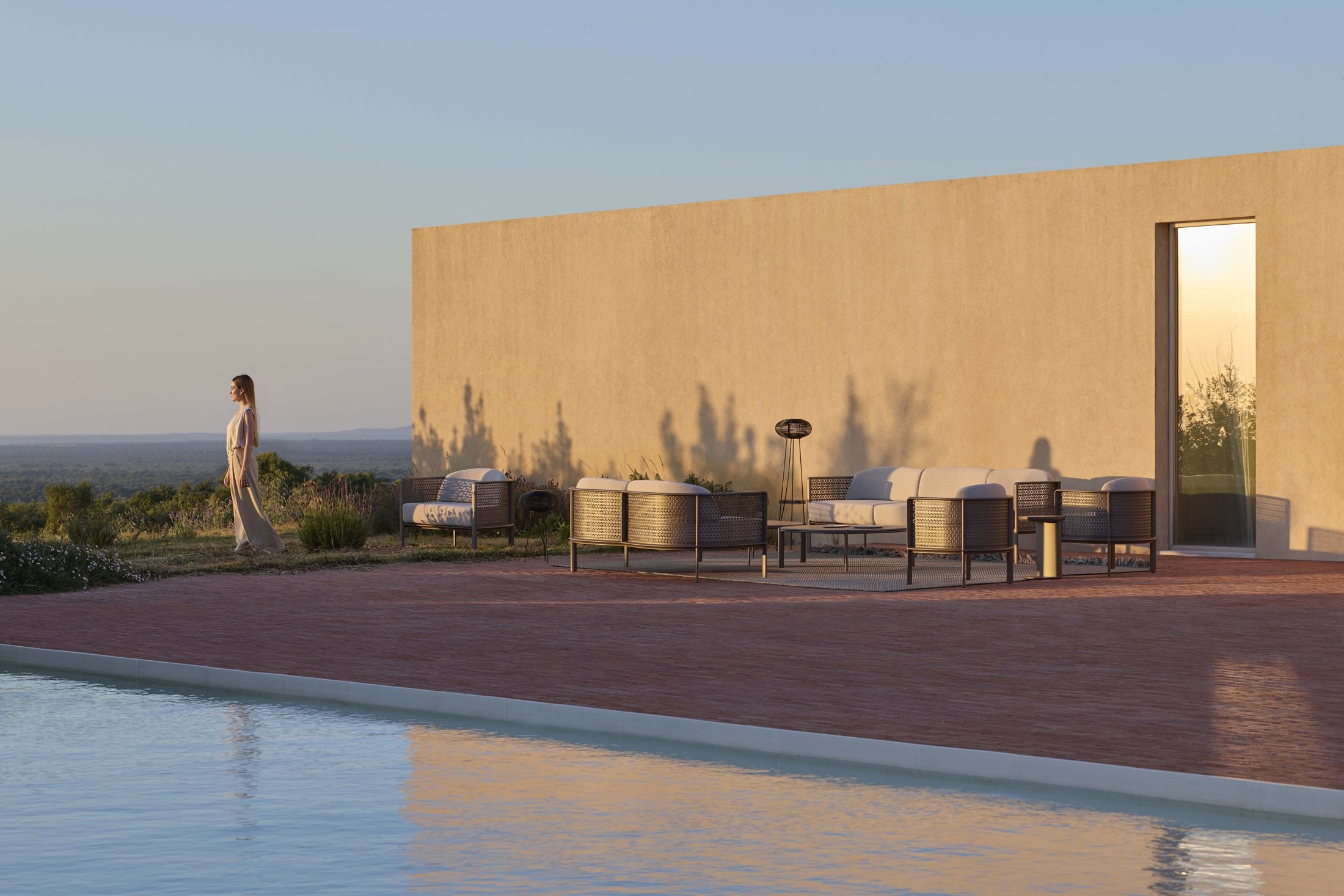 Gandiablasco modular outdoor sofa and armchairs in bronze mesh on a terrace by a pool at sunset, against a plaster wall.